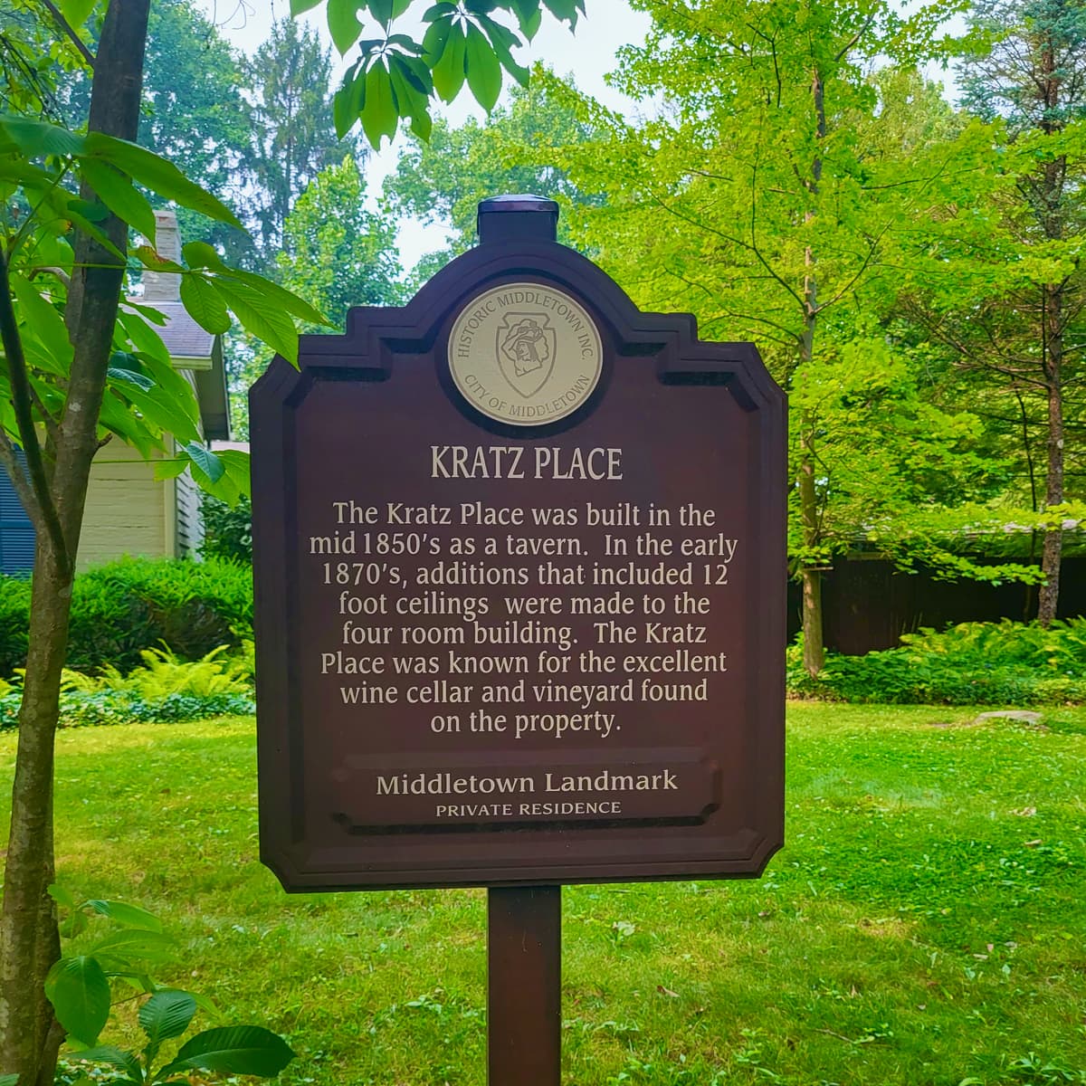 A wooden historical sign for Kratz Place with trees in the background.